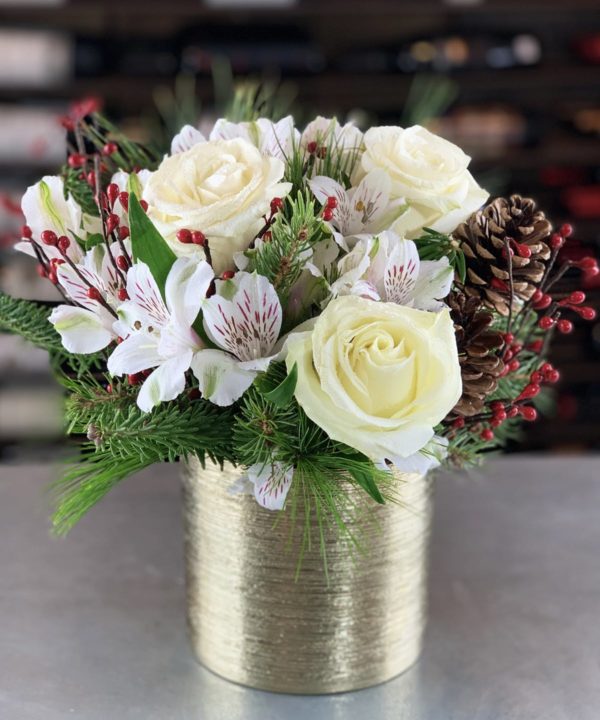 A beautiful keepsake gold cylinder filled to the brim with white holiday greenery, white alstromeria, white winter roses, and red berry accents