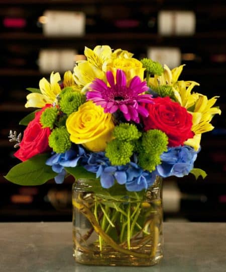 Dazzling and delightful, this bold and colorful flower bouquet is set to impress with its high impact look and graceful styling. Orange & Yellow roses accented with hot pink gerbera daisies and yellow alstroemeria on top of a bed of electric blue hydrangea arranged in a clear cube.